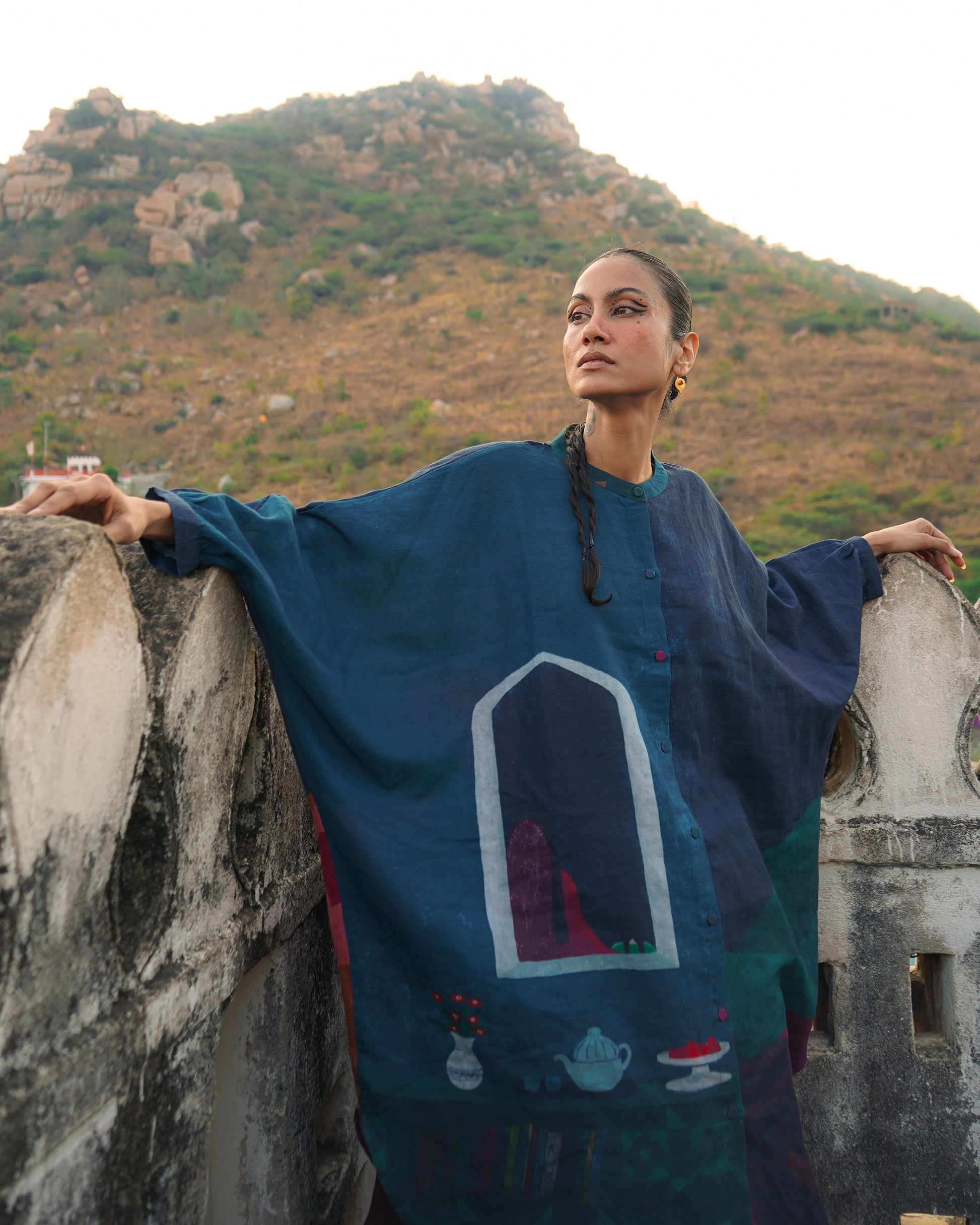 Woman in a blue embroidered dress standing on a stone wall with mountains in the background