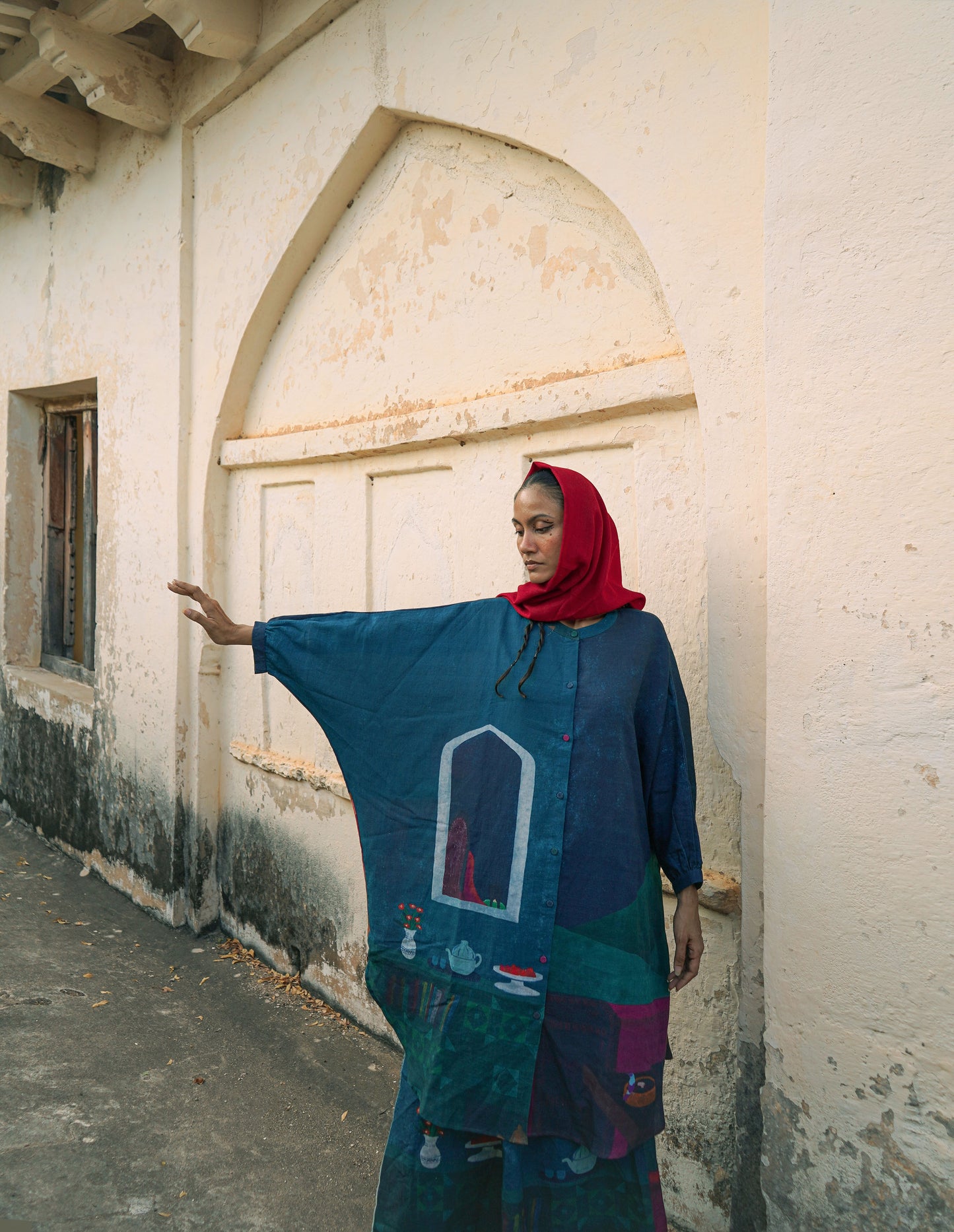 Woman in a colorful dress and red headscarf standing against a white wall.
