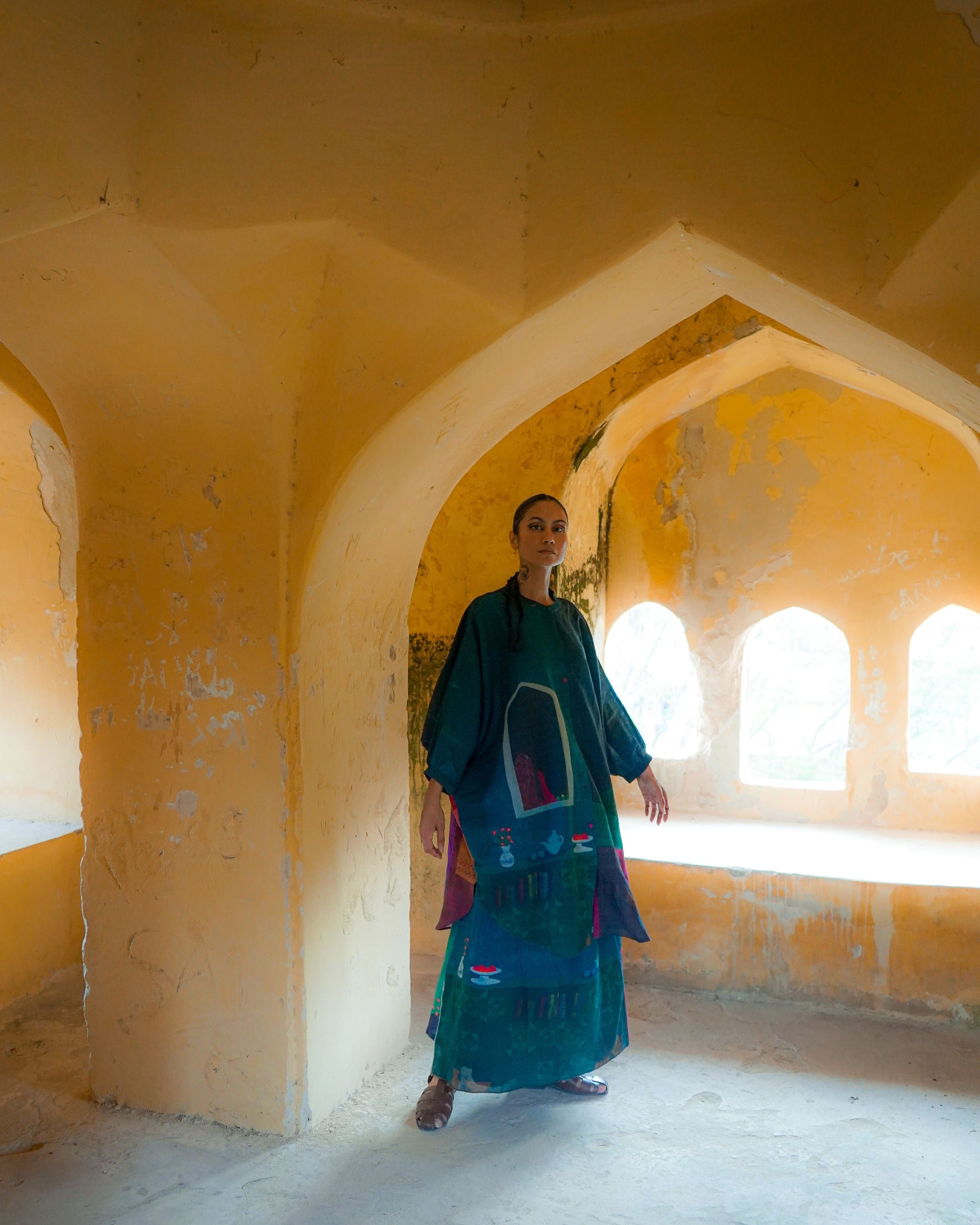 Woman in a green dress standing in a sunlit room with arches and yellow walls.