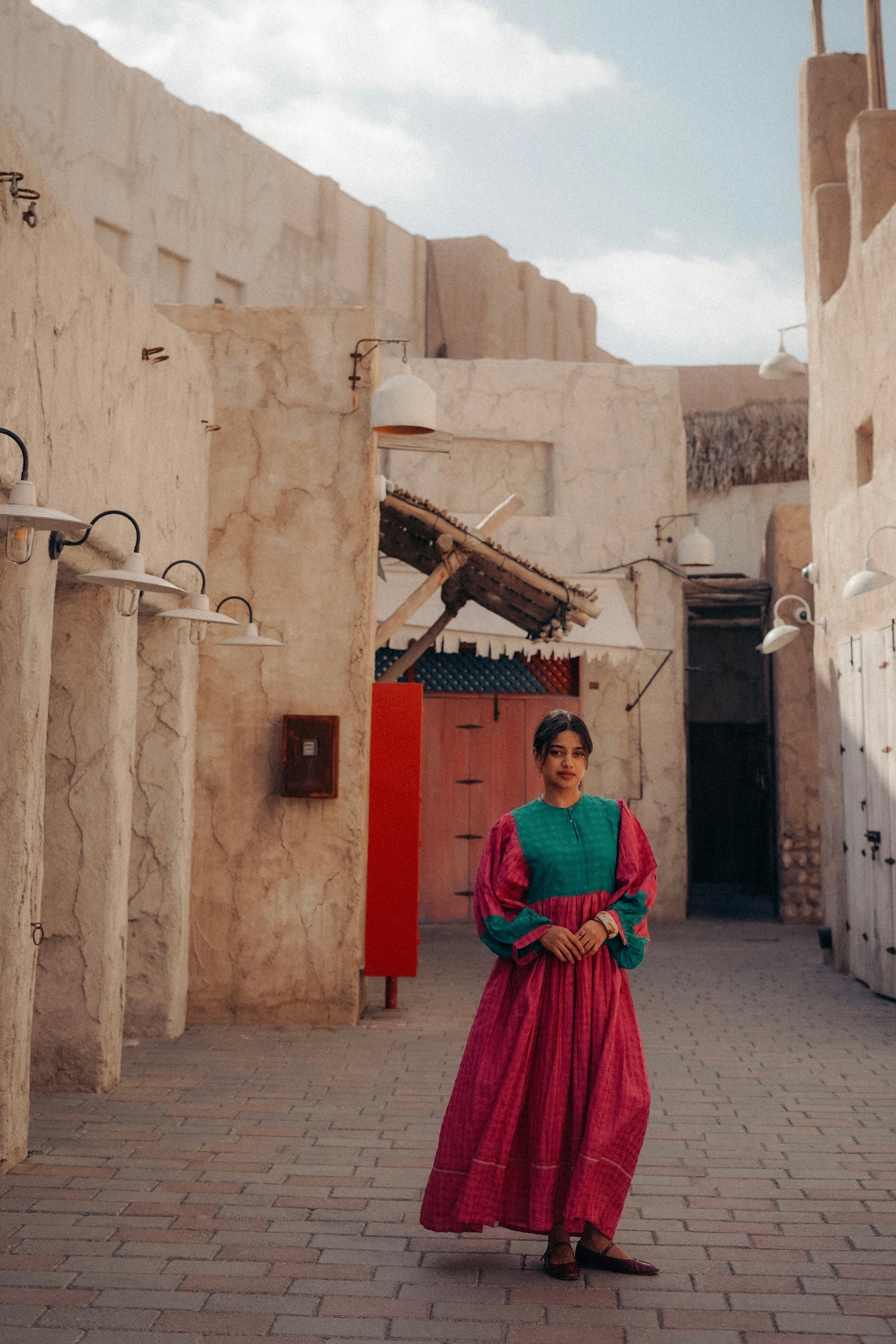 Woman in traditional attire standing in a sunlit alleyway with stone buildings.
