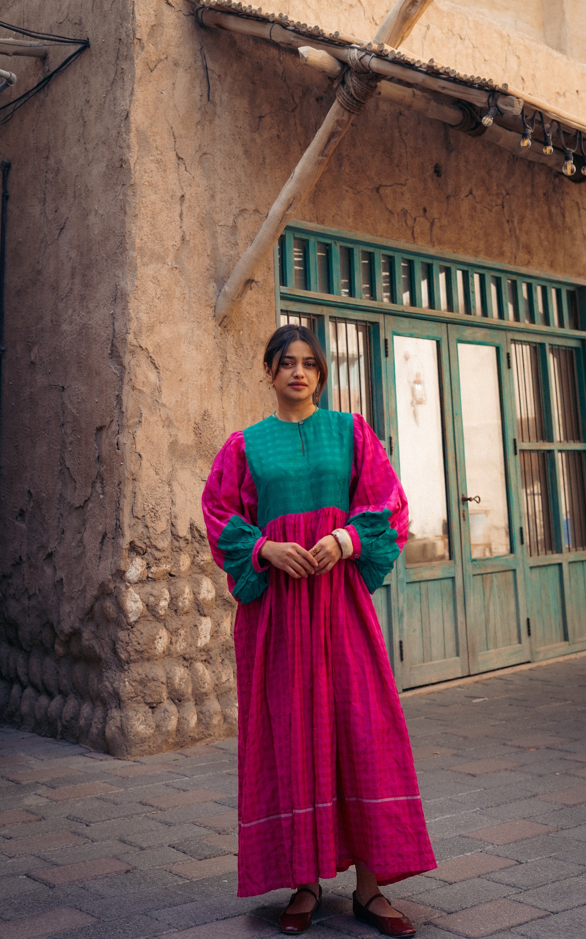 Woman in a pink and green traditional outfit standing in front of a rustic building.