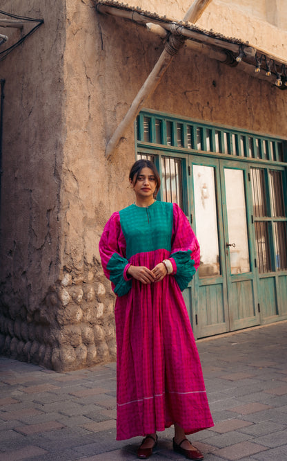 Woman in a pink and green traditional outfit standing in front of a rustic building.