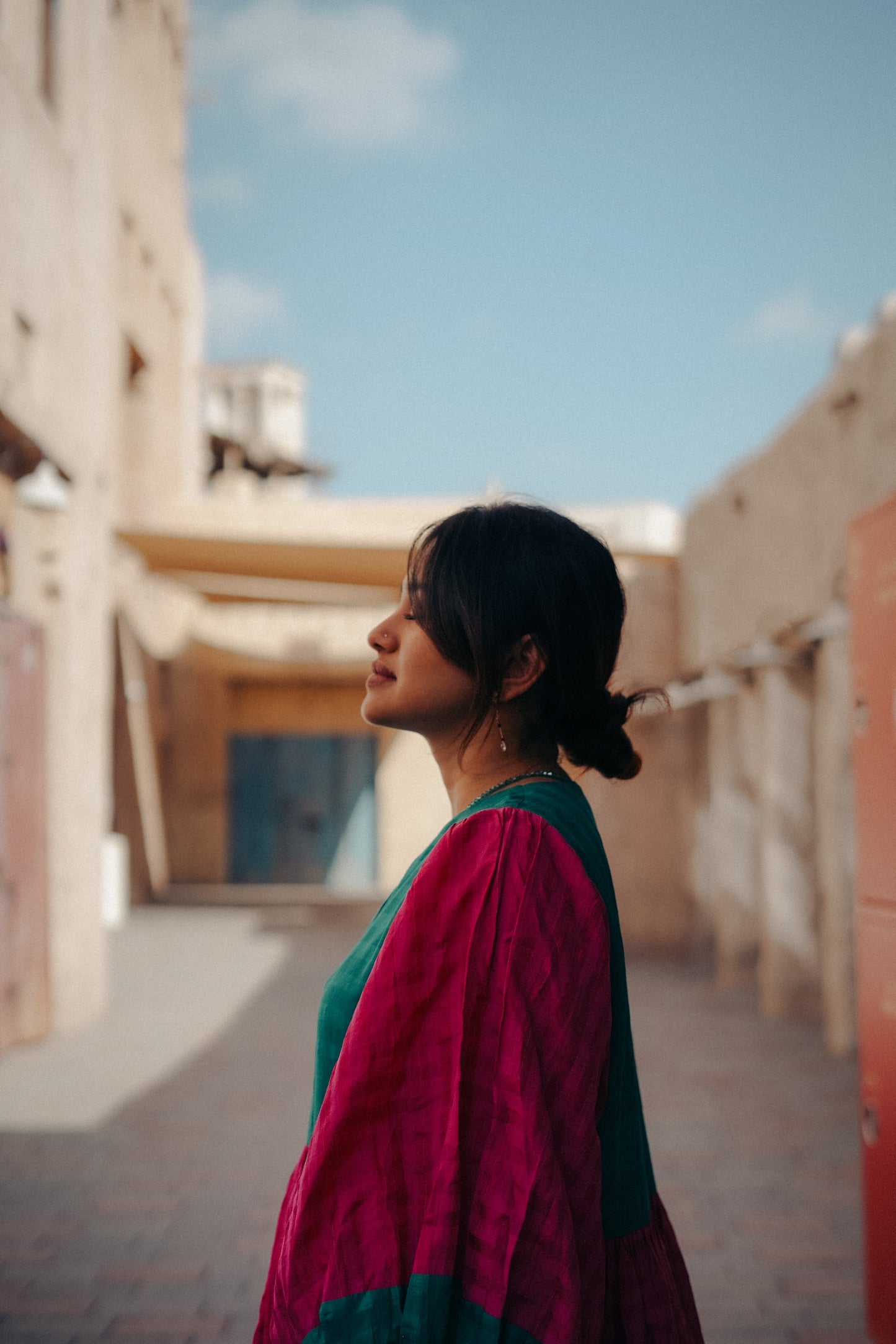 Woman in a pink and green traditional outfit standing in a sunlit alleyway.