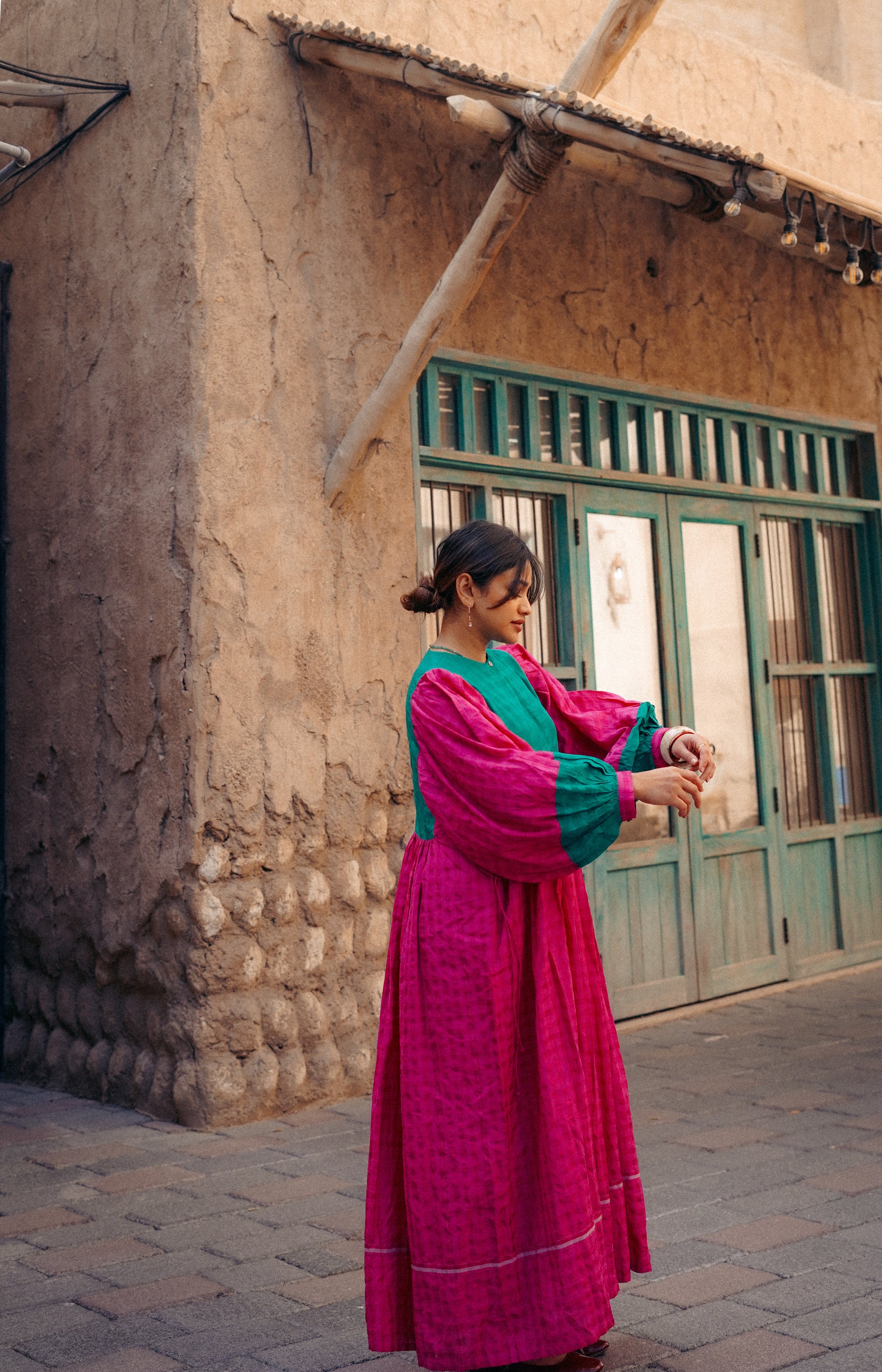 Woman in a vibrant pink and green dress standing in front of a rustic building with a green door.