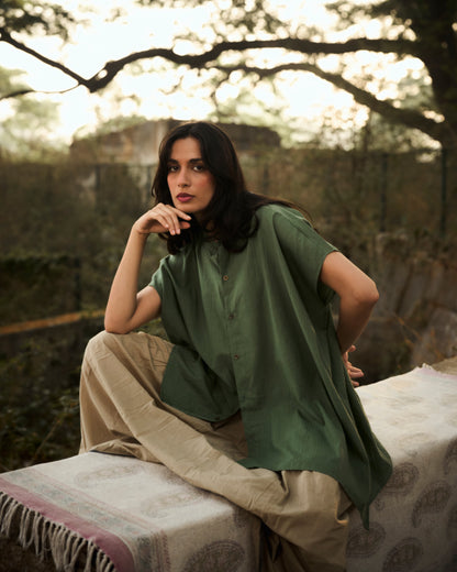 Woman in a green shirt and beige pants sitting outdoors with trees in the background