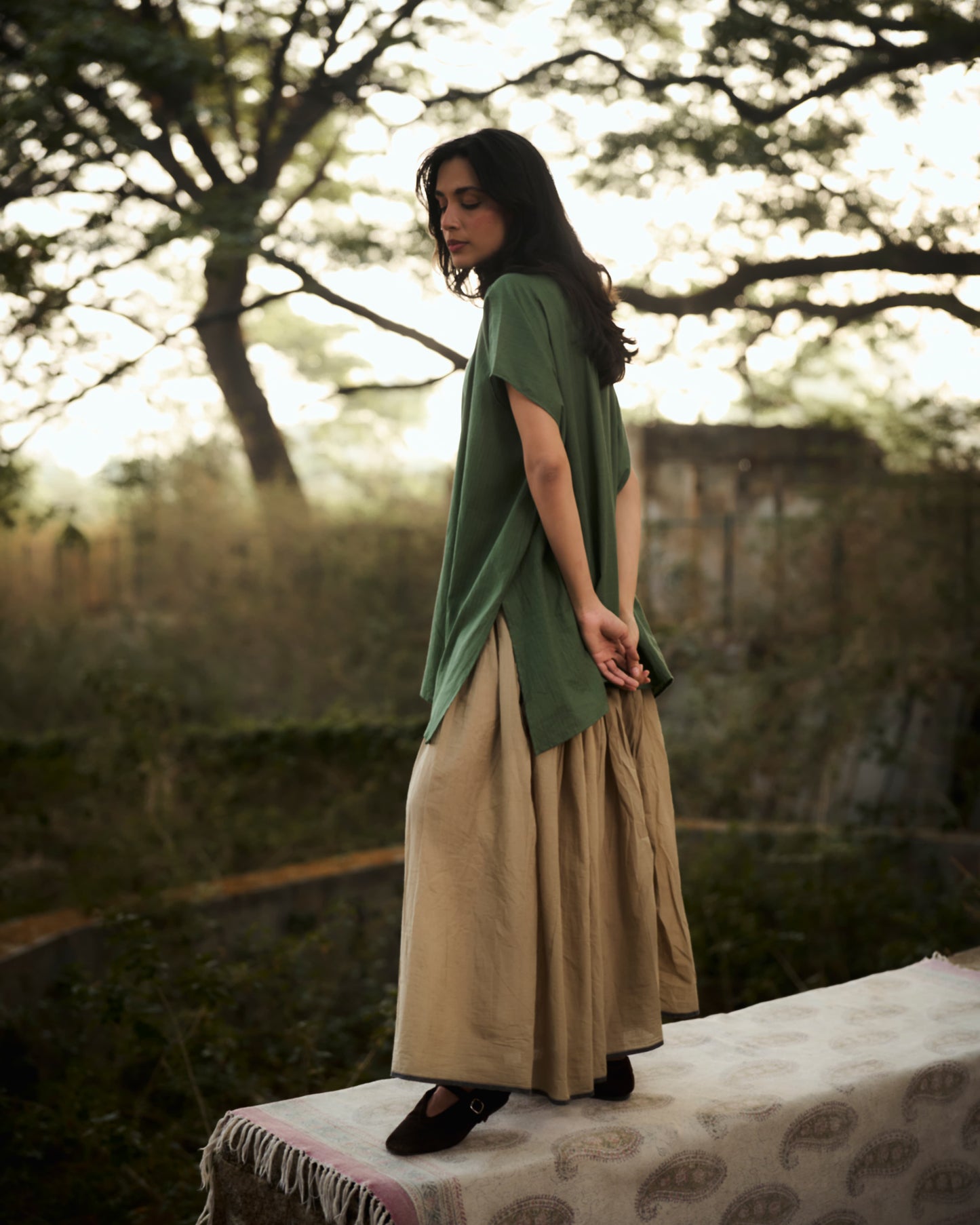 Woman in a green top and beige skirt standing outdoors with trees in the background