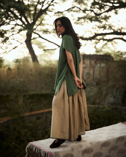 Woman in a green top and beige skirt standing outdoors with trees in the background