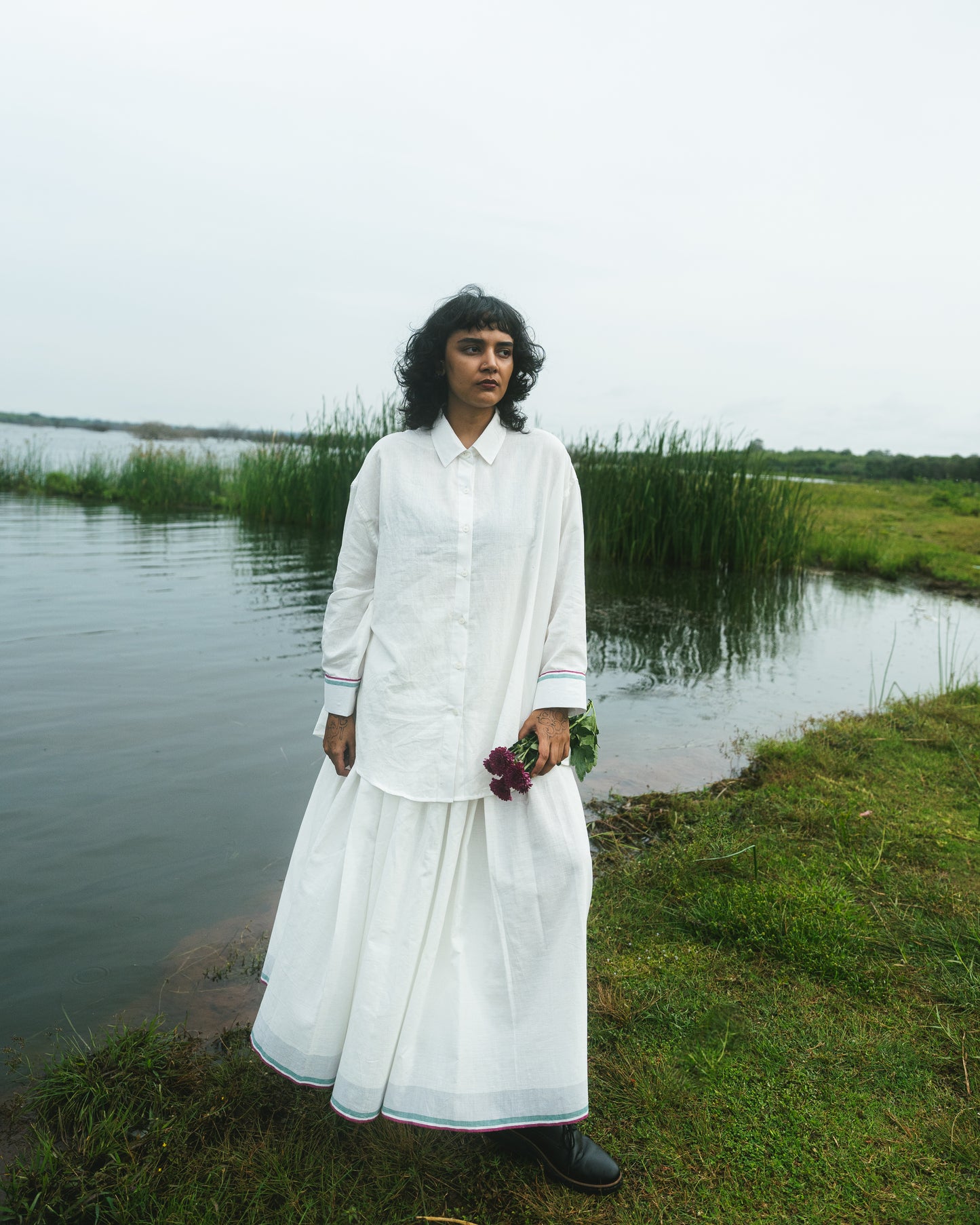 Person in a white outfit standing by a body of water with greenery in the background